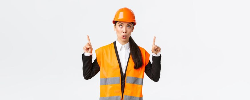 Building, construction and industrial concept. Surprised and curious asian female construction manager, engineer in safety helmet and reflective clothing, pointing fingers up at interesting banner.