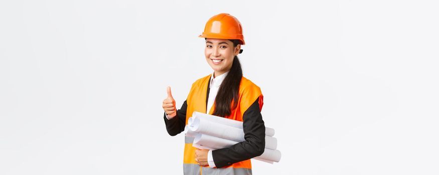 Confident successful female asian construction manager, architect in safety helmet and jacket, showing thumb-up and carry blueprints of building project, guarantee quality, white background.