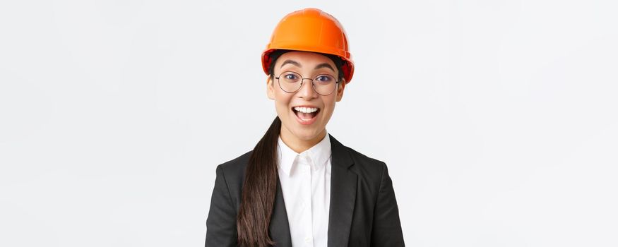 Close-up of excited upbeat asian female chief construction engineer, architect in suit and safety helmet smiling amazed, looking with hope and satisfaction at good work done, white background.