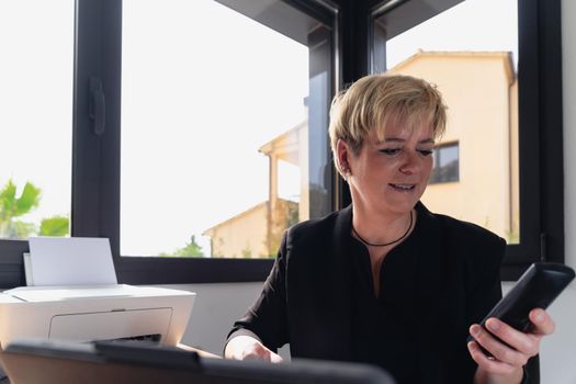 Mature and smiling professional beautician woman with blonde hair, dressed in black work uniform, working in her home office. Owner of a beauty salon receiving phone consultations. Work from home, freelance, business, creative occupation, lifestyle concept. Relaxed atmosphere and soft lighting from window, natural light, work table, desk, computer. Horizontal.