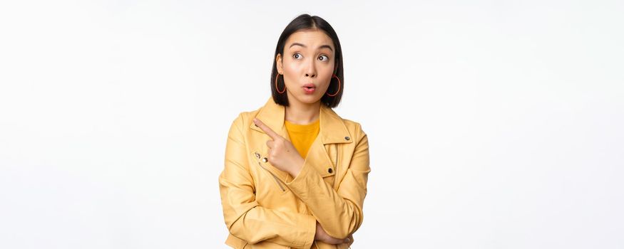 Image of surprised aisan woman pointing finger left, looking at logo or banner with intrigued face expression, standing over white background.