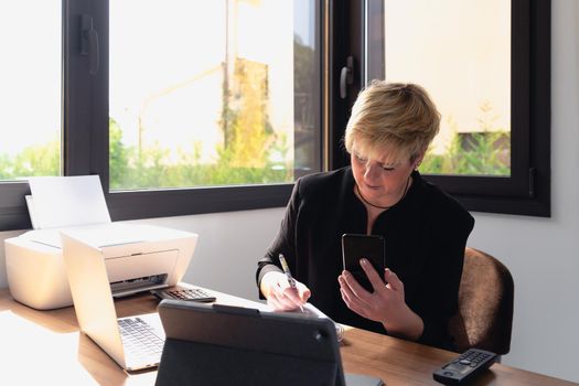 Portrait of a blonde, mature, focused and professional beautician, talking on mobile phone to arrange a visit. She makes a note in the diary of clients' appointments. Owner of a small business Relaxing atmosphere and soft lighting from window, natural light, work table, desk, computer. Horizontal.