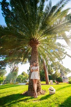 The bright beautiful girl in a light dress and hat standing under a palm tree of Monaco in sunny weather in summer. High quality photo