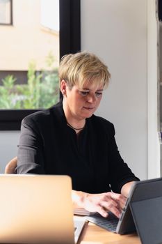 smiling mature professional beautician woman with blonde hair, dressed in black work uniform, working in her home office. secretary of a beauty salon taking appointments on the phone. Work from home, freelance, business, creative occupation, lifestyle concept. Relaxed atmosphere and soft lighting from window, natural light, work table, desk, computer. vertical