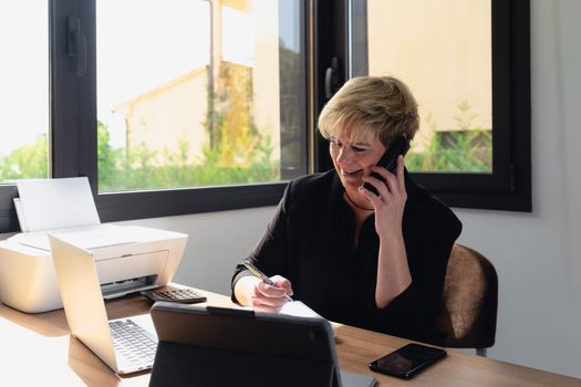 Portrait of a mature, smiling, professional beautician with blonde hair, Mature business woman with blonde hair, working in her home office talking on the phone while consulting her laptop. Small business owner Relaxed atmosphere and soft lighting from window, natural light, work table, desk, computer. Horizontal.