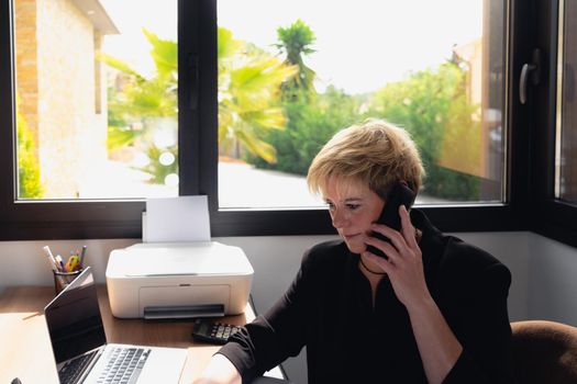 Mature and smiling professional beautician woman with blonde hair, dressed in black work uniform, working in her home office. Owner of a beauty salon receiving phone consultations. Work from home, freelance, business, creative occupation, lifestyle concept. Relaxed atmosphere and soft lighting from window, natural light, work table, desk, computer. Horizontal.