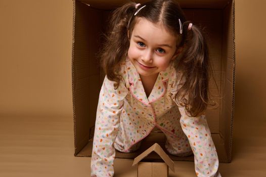 Housing, insurance and real estate concept. Cute child girl peeking out from behind a cardboard box, looking at camera. Craft carton box on a beige background with copy space for ads.