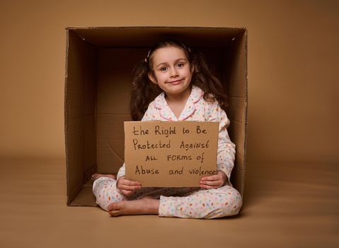 Adorable little Caucasian girl holding a poster with a message to be protected from abuse and violence, sitting inside a cardboard box. Social advertisement for International Children day. 1st July