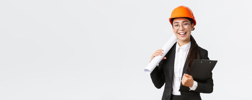 Professional happy asian female architect, construction engineer in helmet and business suit holding blueprints and clipboard with building documentation, smiling cheerful, white background.