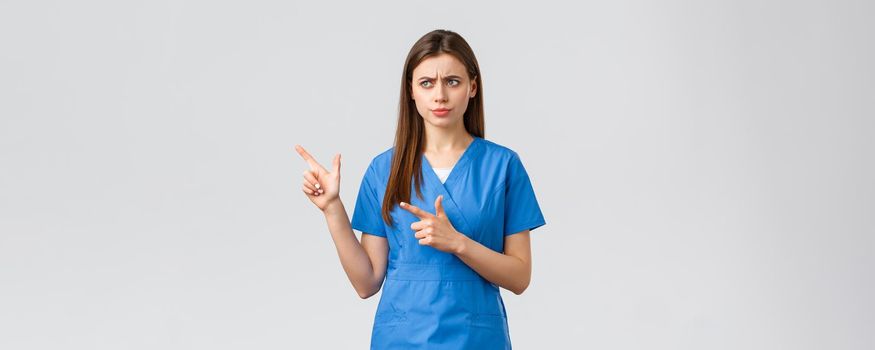 Healthcare workers, prevent virus, insurance and medicine concept. Something wrong. Serious displeased nurse or doctor in blue scrubs, frowning looking and pointing left concerned.