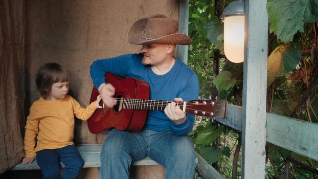 Father cowboy with a small child sits on the porch of an old house and plays an acoustic guitar for his cute baby. The concept of family pastime, relationships and teaching children music