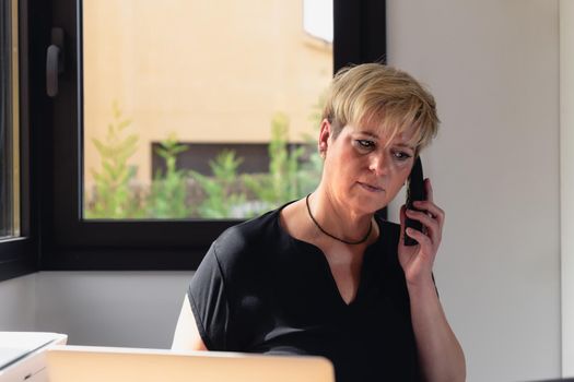 smiling mature professional beautician woman with blonde hair, dressed in black work uniform, working in her home office. secretary of a beauty salon taking appointments on the phone. Work from home, freelance, business, creative occupation, lifestyle concept. Relaxed atmosphere and soft lighting from window, natural light, work table, desk, computer. Horizontal.