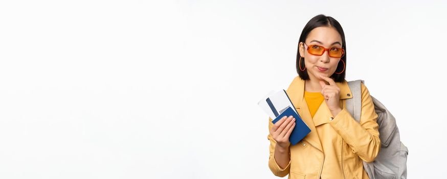 Asian girl tourist with boarding tickets and passport going abroad, holding backpack, thinking of travelling, standing over white background.
