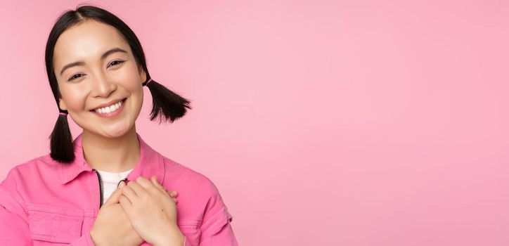 Cute korean girl with silly hairbuns, looking happy and grateful, thank you pose, holding hands on heart flattered, posing against pink background.