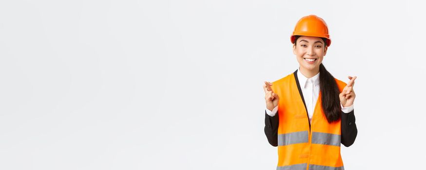 Hopeful and optimistic smiling asian female engineer, construction manager in safety helmet having faith in project, cross fingers good luck and looking positive, standing white background.