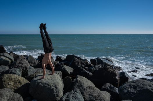 Shirtless man doing handstand on rocks by the sea