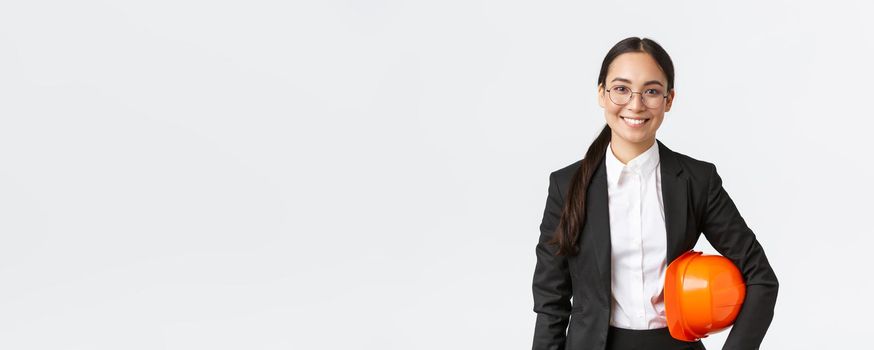 Portrait of happy professiona female asian construction manager, engineer in business suit holding helmet and smiling at camera, entering building area for inspection, white background.