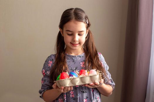 Cute little brunette girl, in cute dress, standing in a room with curtains, holding a cardboard tray with multi-colored hand-painted eggs, looking at them