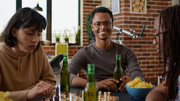 Portrait of african american man having fun with friends in living room, playing chess and board games. Young adult enjoying group gathering to play with beer and snacks for entertainment.