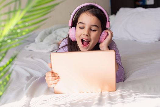 Happy little girl in pink headphones lying on a white bed, holding a pink digital tablet. Copy space