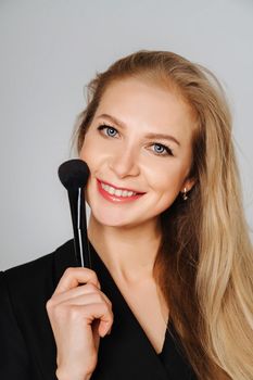 Beautiful middle aged woman makeup waving one makeup brush, winking at the camera and smiling. Blond hair and a black jacket on a light background