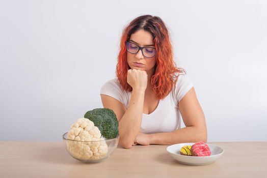 Caucasian woman prefers healthy food. Redhead girl chooses between broccoli and donuts on white background