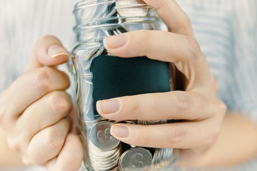 Bottle with coins in women's hands. Blank black sticker on donation money jar. Girl holds her savings. Glass pot for tips.