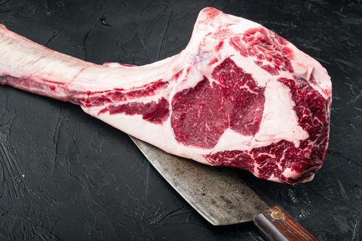 Raw uncooked black angus beef tomahawk steak on bone set, and old butcher cleaver knife, on black stone background, with copy space for text