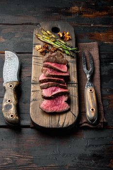Fillet Mignon tenderloin Grilled and sliced meat beef steaks medium rare set, with onion and asparagus, on wooden serving board, with meat knife and fork, on old dark wooden table background