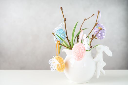 Home interior with easter decor. Vase with willow tree branches with Easter eggs and bunny on white table and background with copy space.