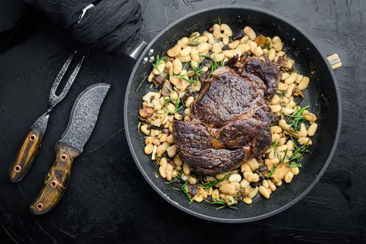 Rib eye steak with herbs, roasted beans, thyme and garlic set, on frying cast iron pan, with meat knife and fork, on black stone background, top view flat lay