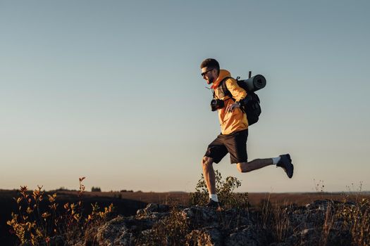 Travel Man Dressed in Windbreaker with Backpack and Camping Mat, Jumping on Top of Hill During Sunset, Male Nomad with Digital Camera Enjoy His Best Solo Trip