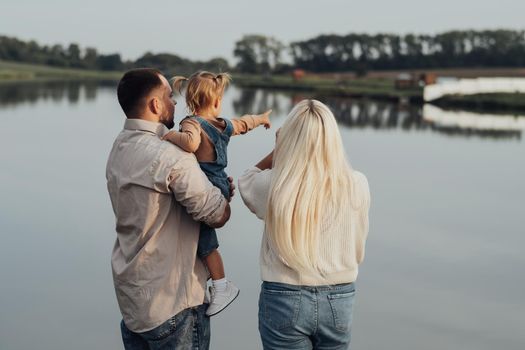 Back View of Young Family Standing Outdoors, Father Holding His Child in Hands, Little Daughter Showing Lake to Her Dad and Mom