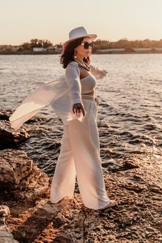 A woman in a white pantsuit and hat is standing on the beach enjoying the sea. Happy summer holidays.