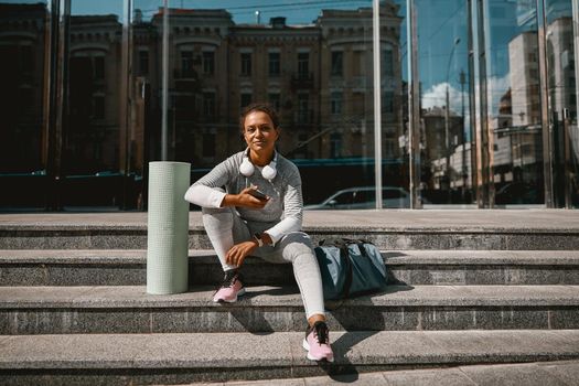 Sporty woman with mat and sports bag sitting after training outdoors. Active lifestyle concept