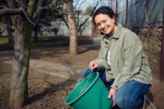 Middle aged Caucasian woman gardener smiles while watering plants from bucket in the garden of her country house. Horticulture, agriculture, Earth Day, home gardening concept