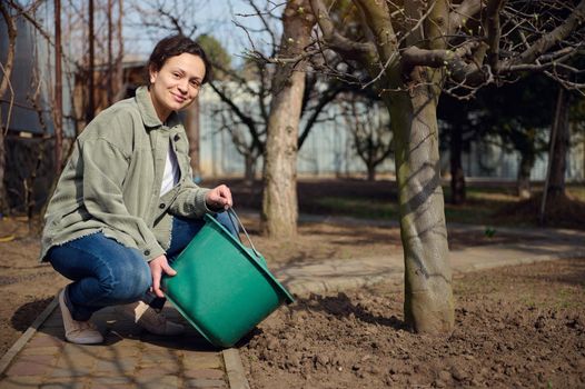 Beautiful smiling woman gardener waters tree from a bucket, enjoying gardening on beautiful sunny spring day. Springtime,horticulture, hobby, Earth protection day, growth of eco vegetables and fruits