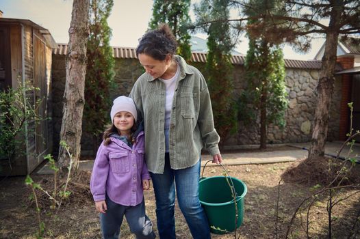 Young pleasant woman holding a bucket with water, standing with her cute daughter and watering baby plants, enjoying gardening in the garden of a backyard of a country side house on n early spring day