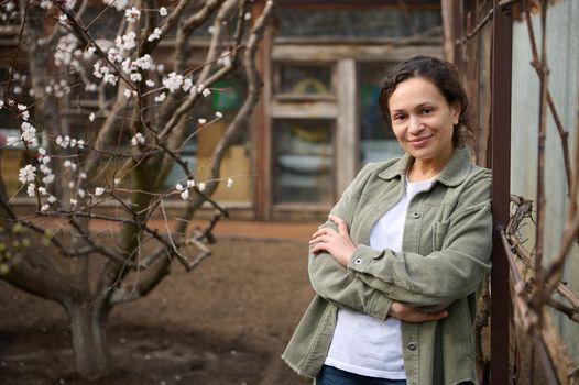 Confident mature woman gardener enjoying an early spring day standing in her botany garden against the flowering fruit trees background