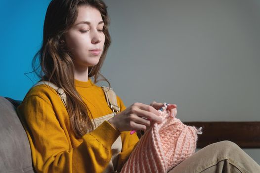 Young happy caucasian woman smiling sitting on sofa and crocheting wool product. Women's hobby production of clothes from wool.