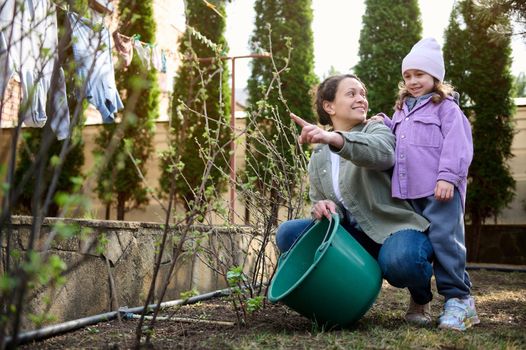 Happy mother and her adorable daughter, cute child girl in the garden, watering blooming plants on an early spring day. Earth day. Environment conservation, ecology concept