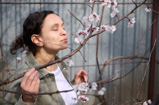 Close-up dark-haired woman enjoying the scent of the white flowers on the branch of a blossoming apricot fruit tree, standing in her garden plot on an early spring day