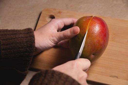 Close-up of woman's hands cutting fresh mango on wooden cutting board at home kitchen.