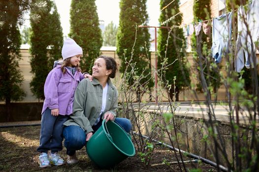 Happy mother and daughter watering flowering bushes and plants in the garden of the backyard of a country house, against the background of drying clothes on a rope on an early spring day