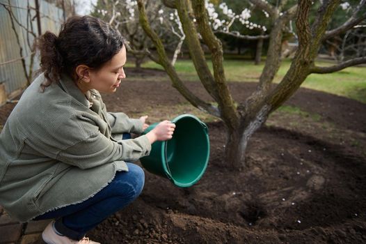 Serene beautiful woman gardener watering a fruit tree from a metal bucket in the garden of her country house.