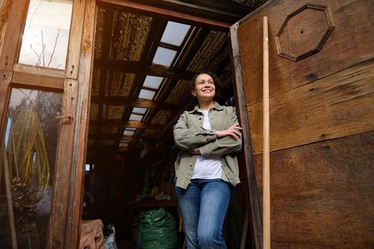 View from the bottom to a serene confident female gardener , cheerful woman standing with crossed arms in her wooden countryside workshop with gardening tools and smiling looking at camera