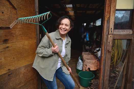 Happy pleasant mature female gardener holding a rake and smiling with a cheerful toothy smile while standing in her wooden rural workshop with gardening tools enjoying home gardening on a spring day