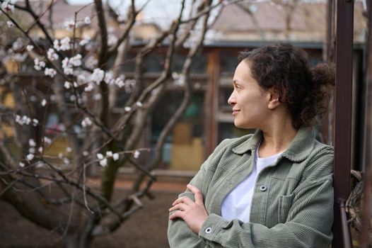 Confident middle aged Caucasian woman gardener enjoying an early spring day standing in her garden plot against the blossoming apricot fruit trees background