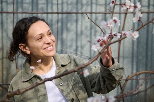 Pleasant multi-ethnic middle aged woman smiles with cheerful toothy smile, enjoying a sunny early spring day standing near a blossoming apricot fruit tree