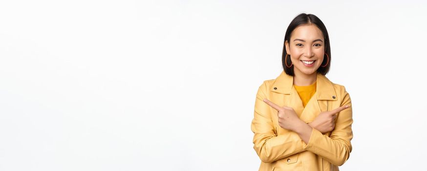 Image of indecisive asian girl, pointing fingers sideways, pointing left and right, choosing variant, deciding, standing over white background. Copy space